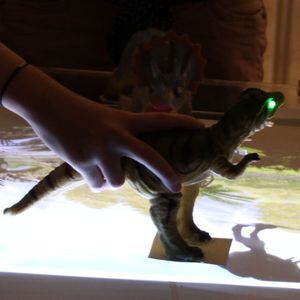 Niño colocando un dinosaurio sobre la mesa proyectora de imagenes y animaciones que luego de ser detectado por el sistema enciende sus ojos de color verde.