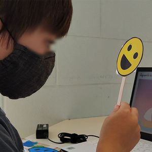 Criança interagindo com o emoji feliz (amarelo) com código QR e olhando o feedback do sistema Aquarela Virtual na tela do computador