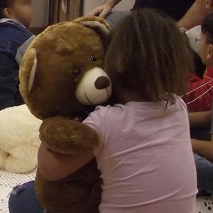 Crianças sentadas no chão abraçando e passando o Teddy, um urso de pelúcia com tecnología embutida, de mão em mão.