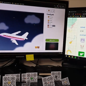 Investigador probando funcionalidades del sistema "Aquarela virtual", con dos monitores que muestran diferentes partes del sistema y una colección de códigos QR impresos colocados sobre el teclado.