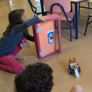 Um garoto está criando um programa tangível, uma garota está segurando um objeto em forma de porta e o outro garoto está observando o movimento do robô mBot.